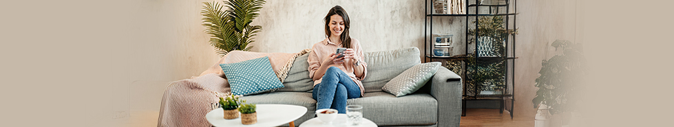 Uśmiechnięta kobieta siedzi na kanapie w mieszkaniu, trzyma w dłoniach telefon i patrzy w ekran.