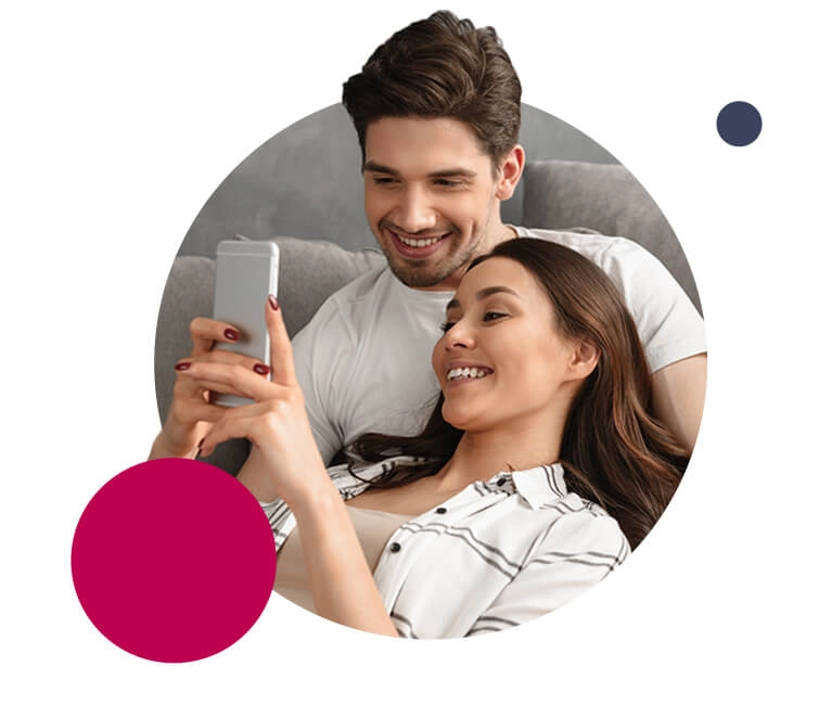 Szczęsliwa i uśmiechnięta para siedzi na kanapie. Oboje patrzą w telefon, który dziewczyna trzyma w ręku