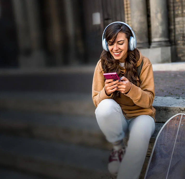 Osoba siedzi na deskorolce na brukowanej uliczce między kamiennymi budynkami i korzysta ze smartfona. Na głowie ma duże słuchawki, a obok widoczna jest grafika promocyjna z napisem „DO 700 zł”.